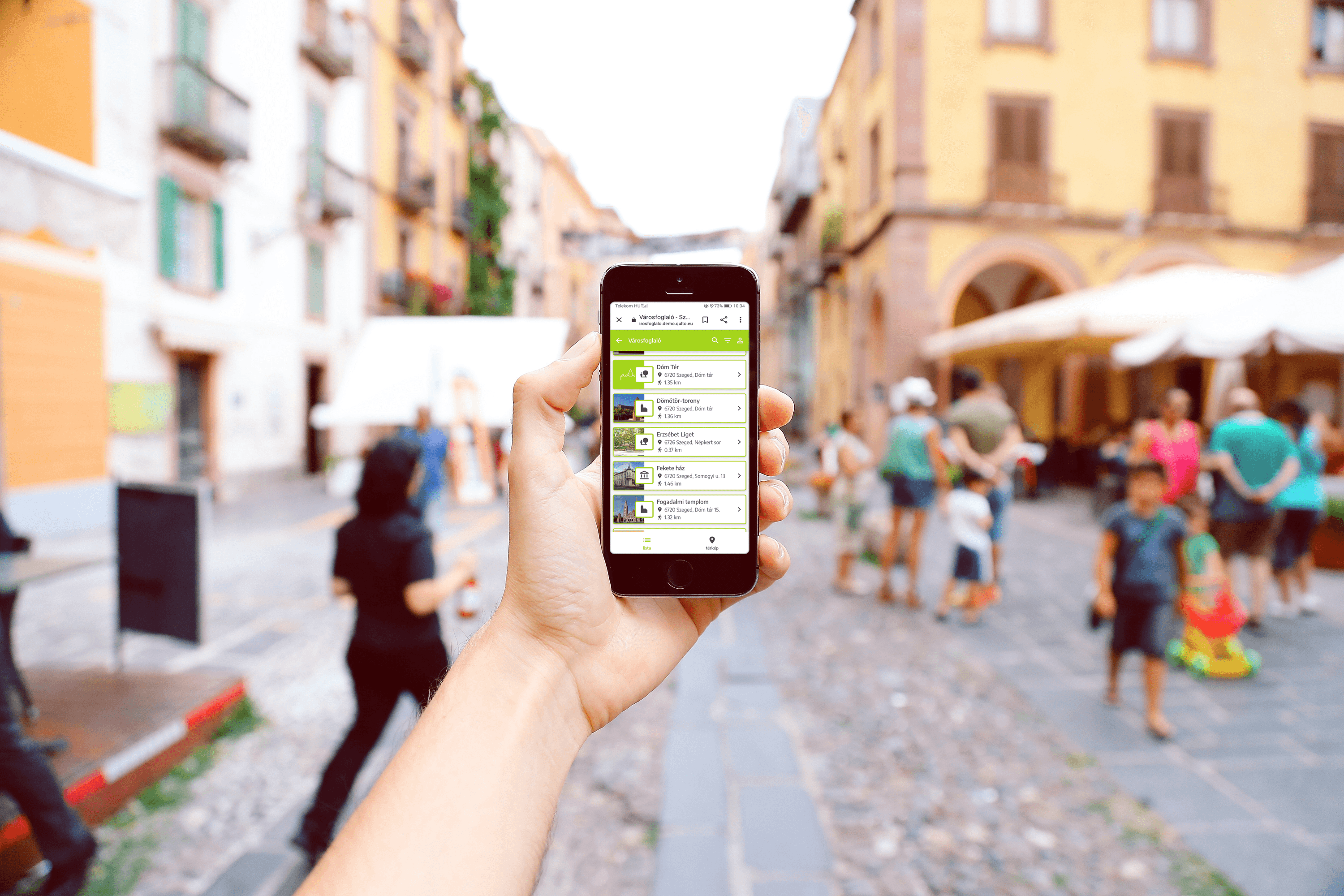 A hand holding a phone showing the Városfoglaló list of sights in Szeged, with a busy old-town street behind it